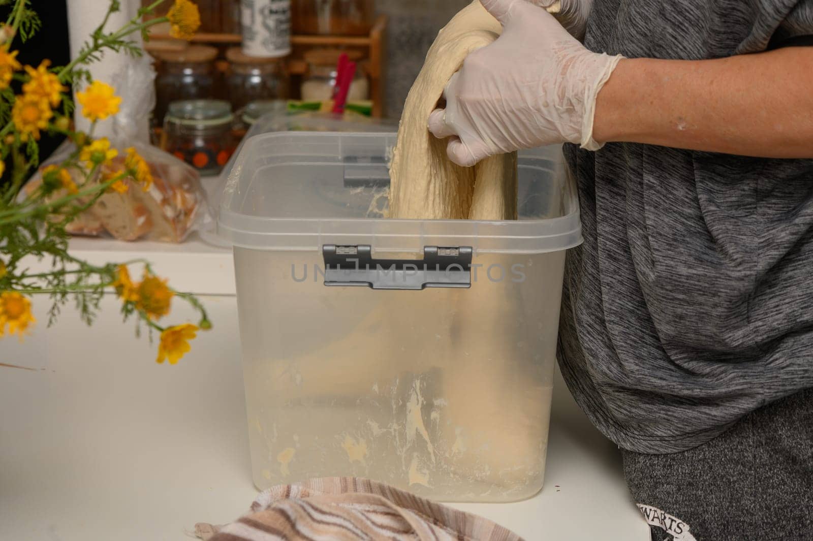 Hands skillfully stretch fresh dough inside a clear container on a kitchen counter adorned with vibrant flowers. The warm sunlight creates a welcoming atmosphere.