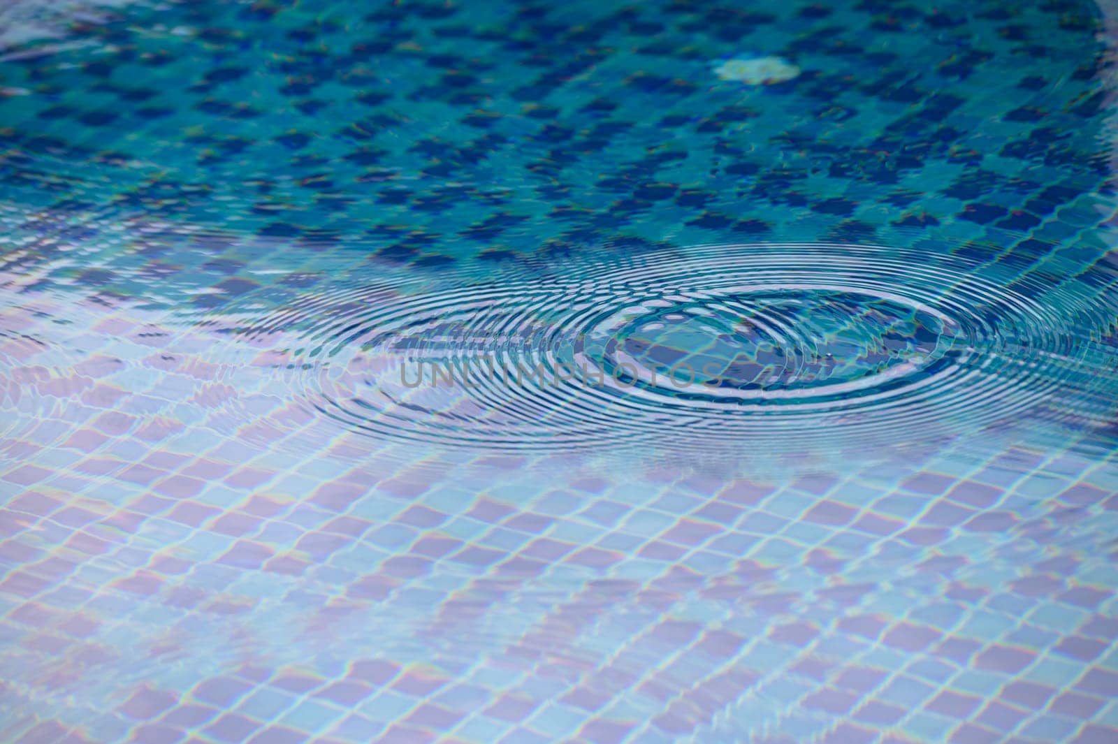 Gentle ripples expand across the shimmering surface of a serene pool, reflecting the sunlight and showcasing intricately arranged blue tiles. A peaceful moment unfolds in the warmth of the day.