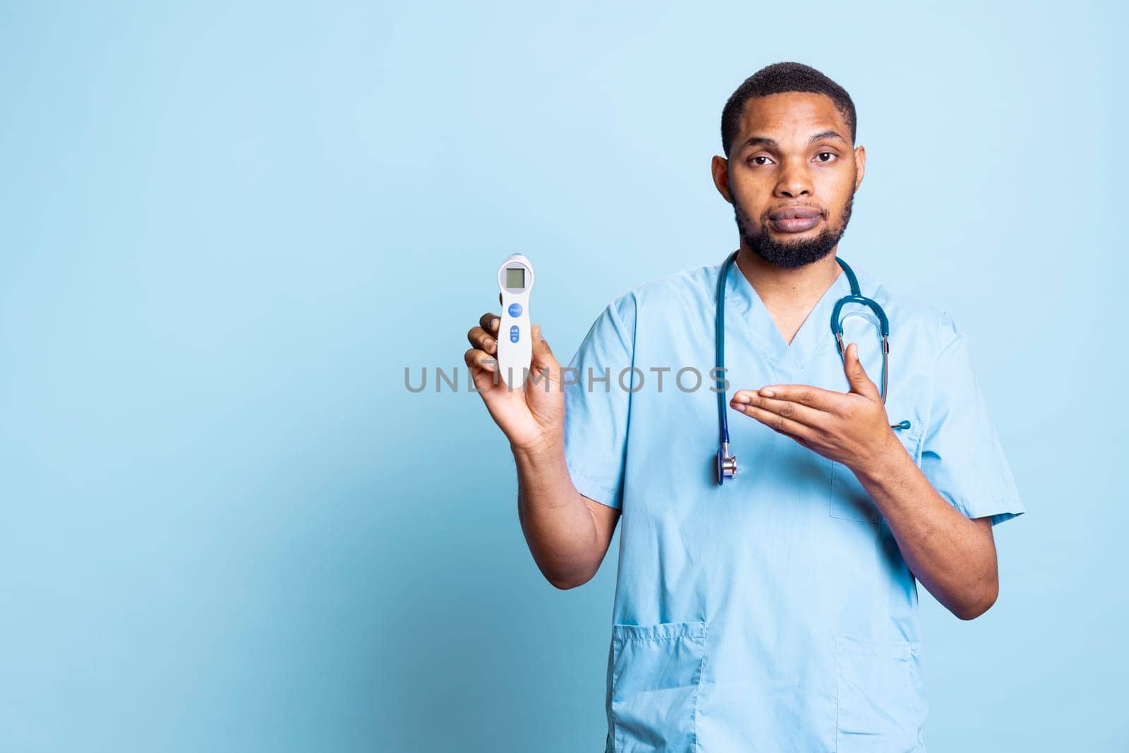 African american medical assistant presenting the digital thermometer against blue background, using tool for fever measurement and to combat virus. Flu symptoms.
