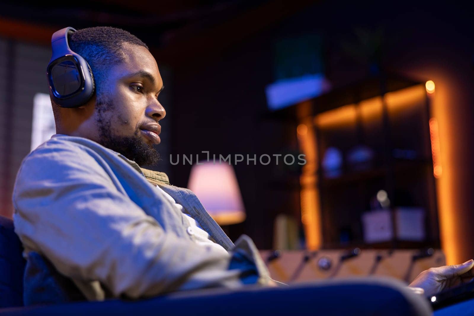African american freelancer listening to a podcast and being focused on online work, using headphones while seated on the couch in his living room. Multitasking in a casual home.