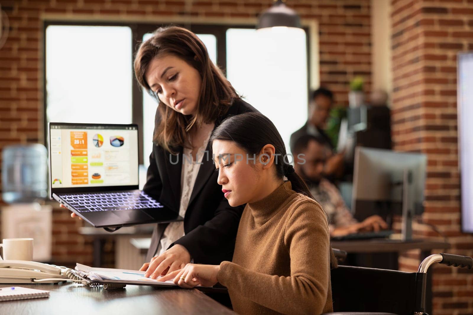 Asian employee at desk participates in business discussions while managing her recovery in modern office. Businesswoman stands with laptop, presenting marketing insights to colleague in wheelchair.