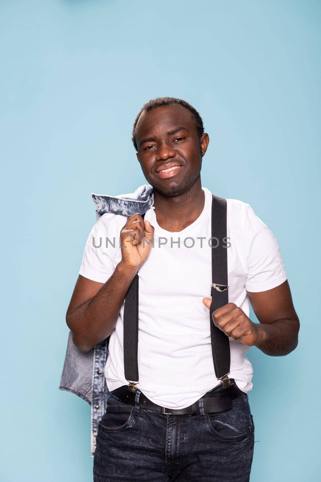 African american man smiles at the camera, standing and holding his denim jacket over his shoulder. Black male model posing against an isolated background, gripping his suspenders.
