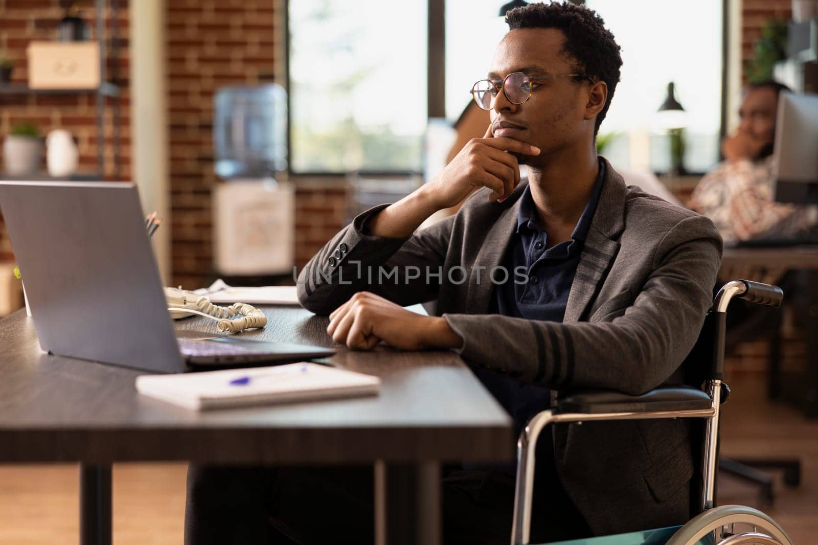 Black businessman in wheelchair holding his chin and concentrating on financial data on his laptop. Male employee with a disability, focused on reviewing a business project plan in startup office.