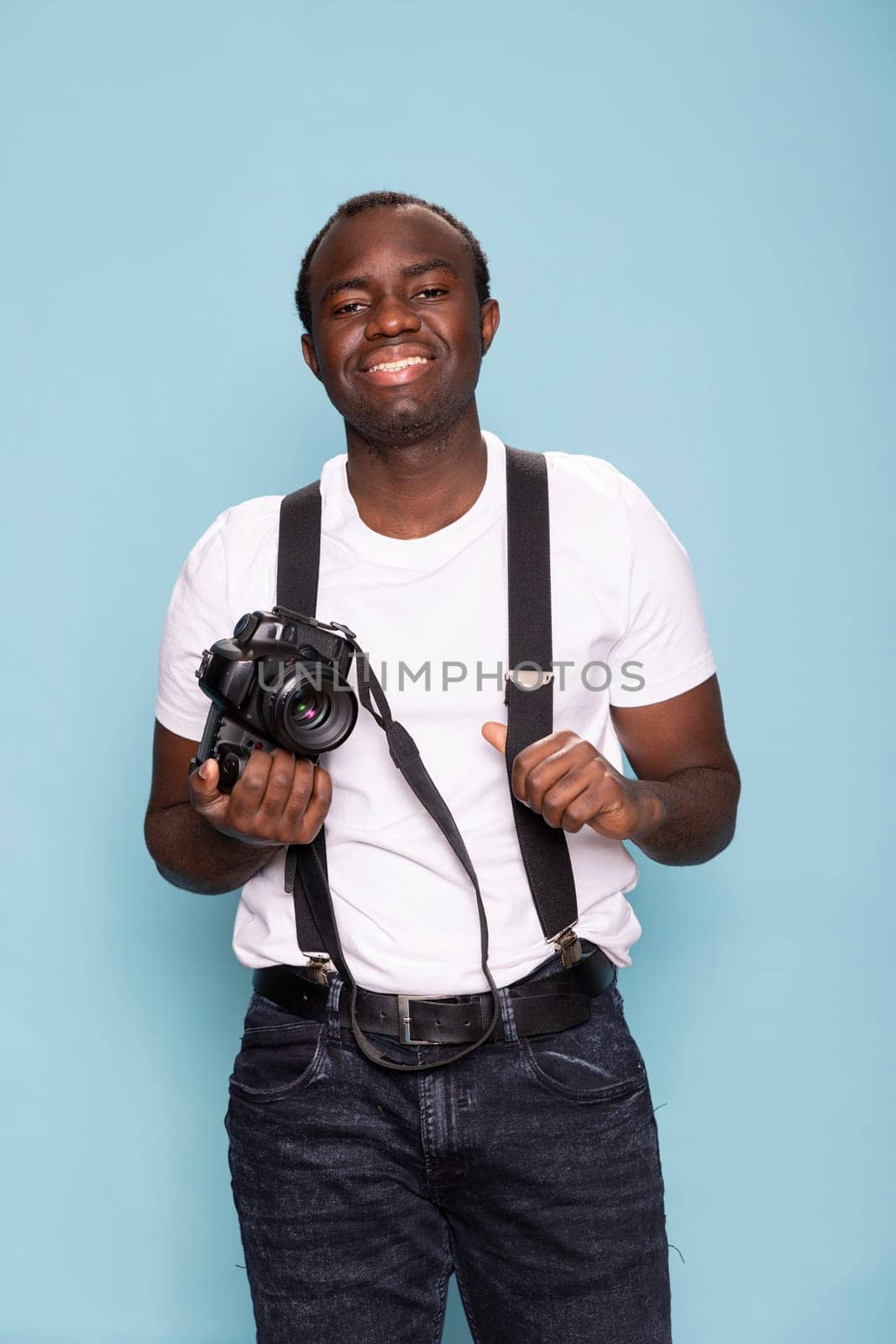 Young man in suspenders and a casual white t shirt holding a professional camera in blue background studio. His warm smile and relaxed posture conveys a welcoming and engaging presence.
