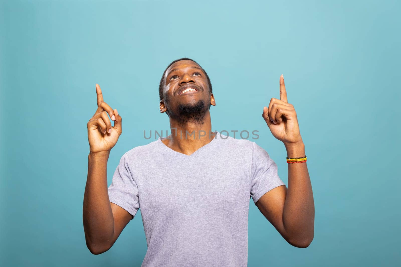 Cheerful african american man pointing upward, looking above as if highlighting a message. Happy male individual in a casual t shirt against a blue background, perfect for an advertisement.