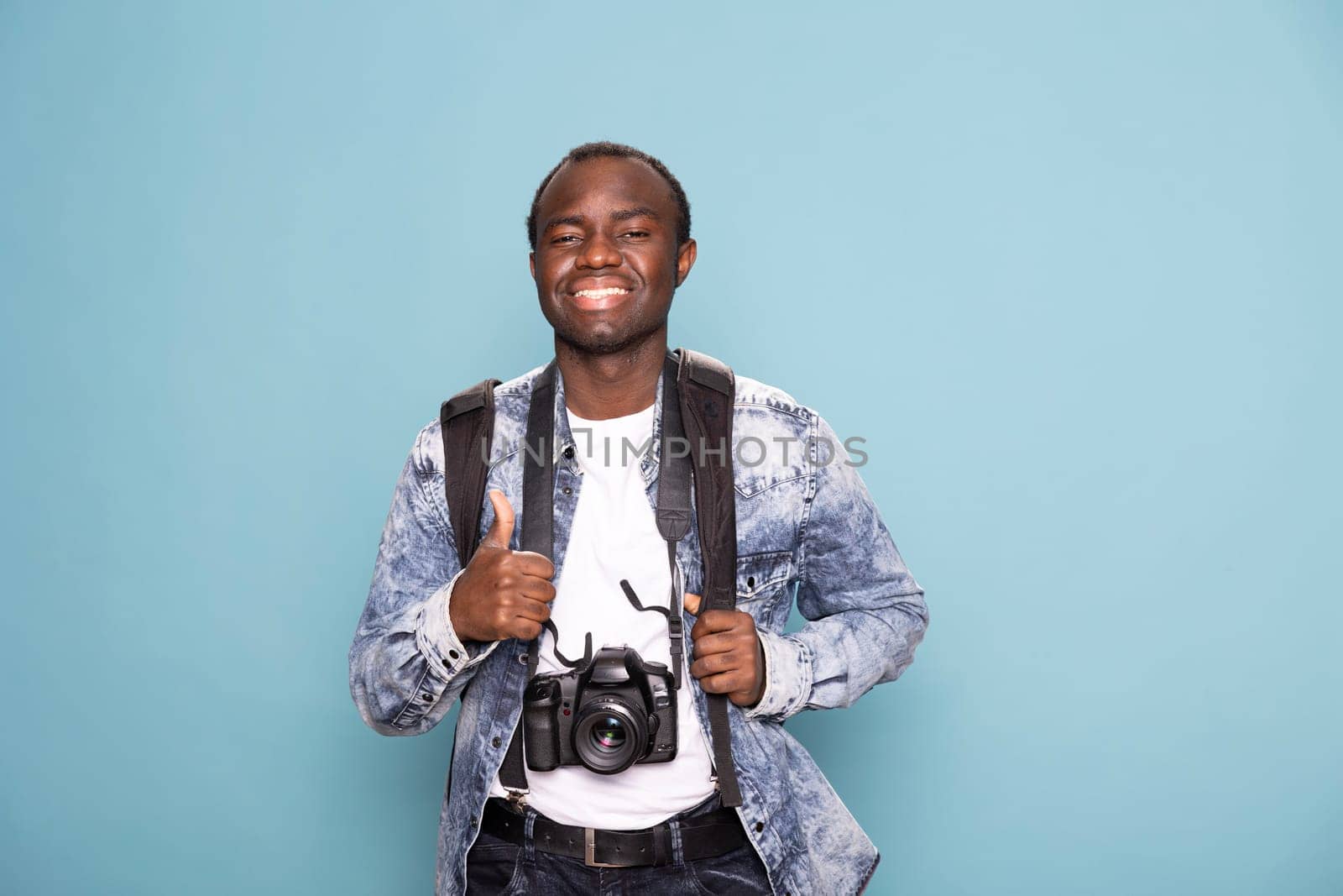 Cheerful black photographer enthusiastically gestures a thumbs up, ready for his next project. African american cameraman smiles brightly, equipped with a backpack and DSLR camera.