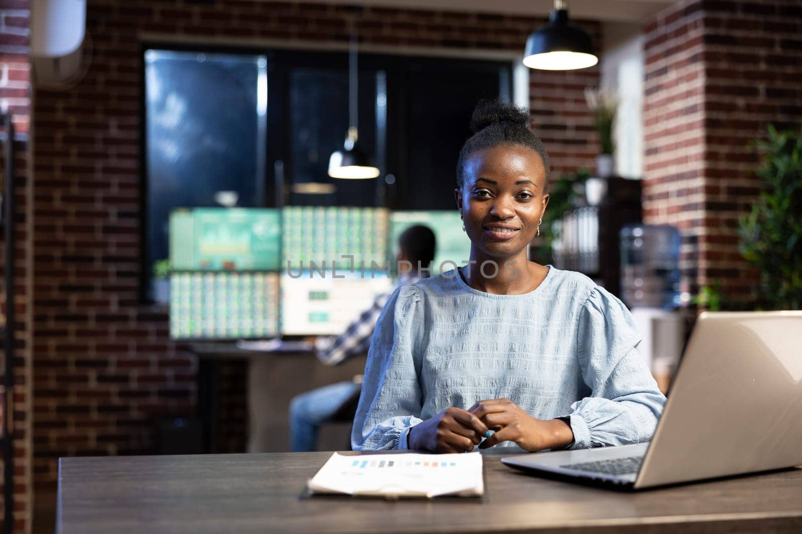 Black woman working late at night in modern office, using her laptop and clipboard at her desk. African american female stock analyst sitting at table and looking at camera, ready for her work shift.