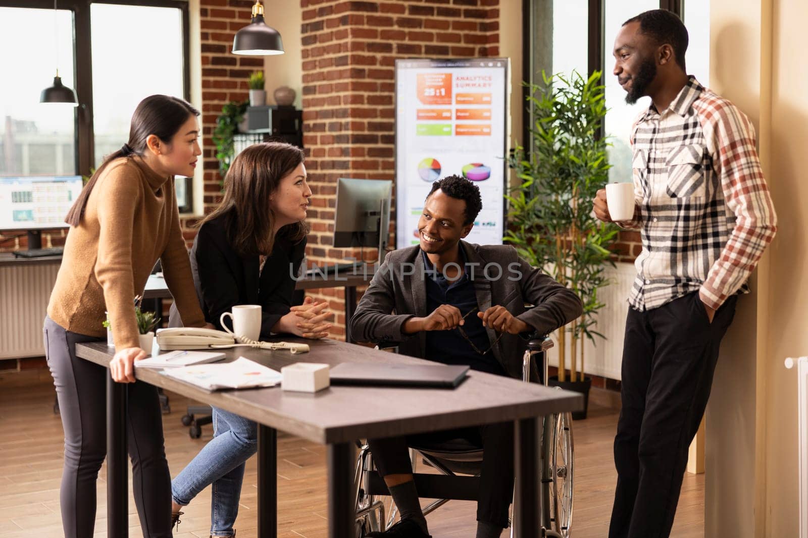 African american wheelchair user and his coworkers, listening intently to caucasian woman share a hilarious story. Male and female employees enjoying a break in brick wall office.