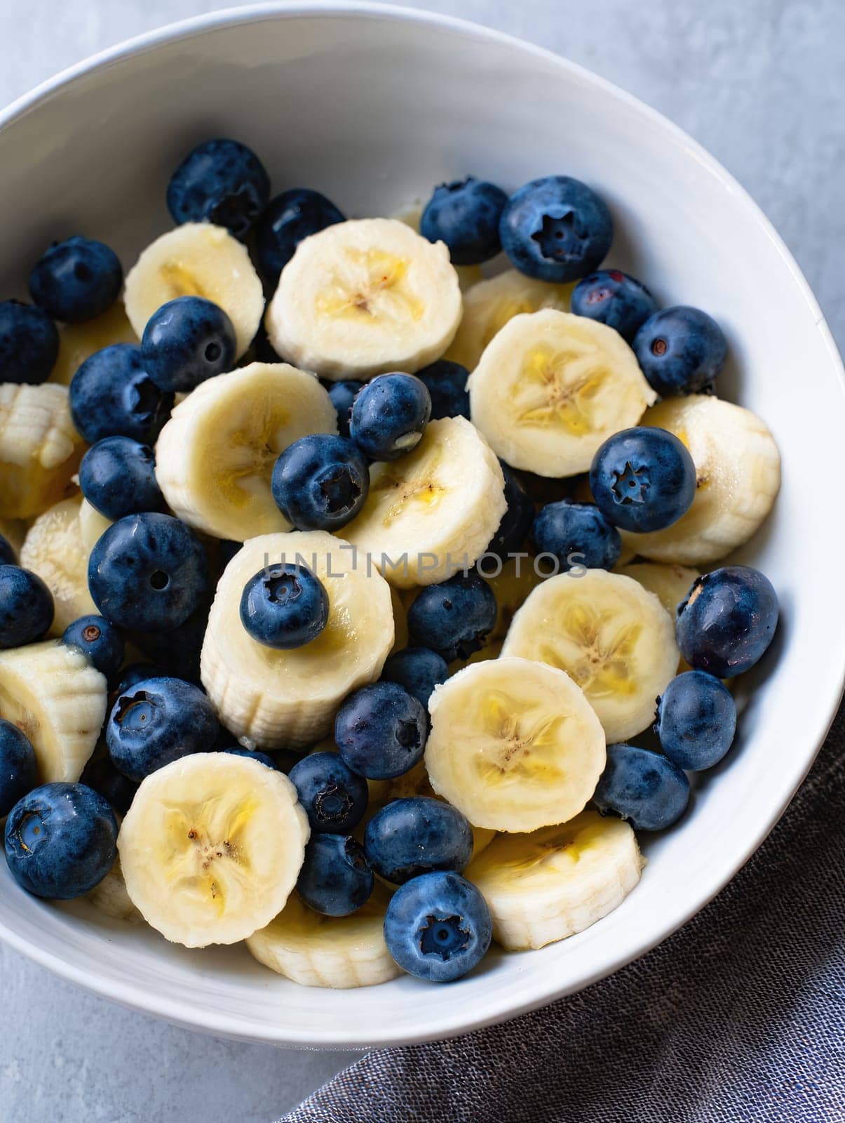 A bowl filled with sliced bananas and fresh blueberries sits on a gray surface. This nutritious combination offers a vibrant look and a delicious flavor, perfect for a healthy snack.