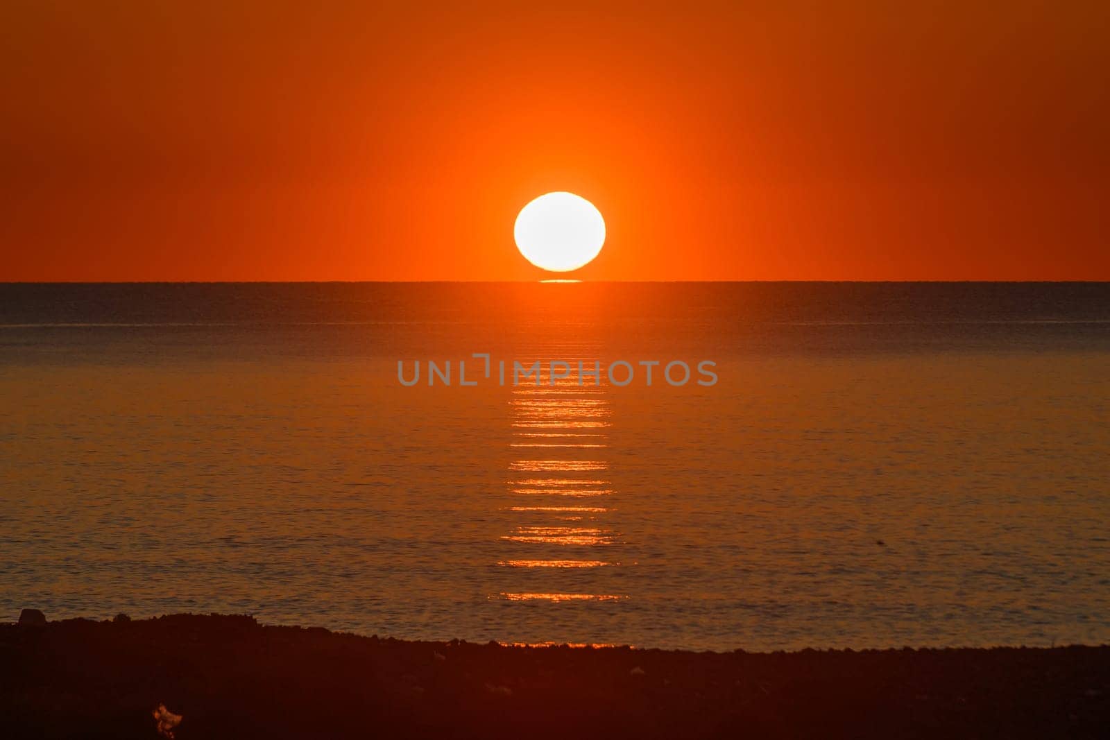 Vibrant hues of orange and gold paint the sky as the sun dips below the horizon, casting shimmering reflections on the calm ocean surface, creating a tranquil and magical atmosphere.