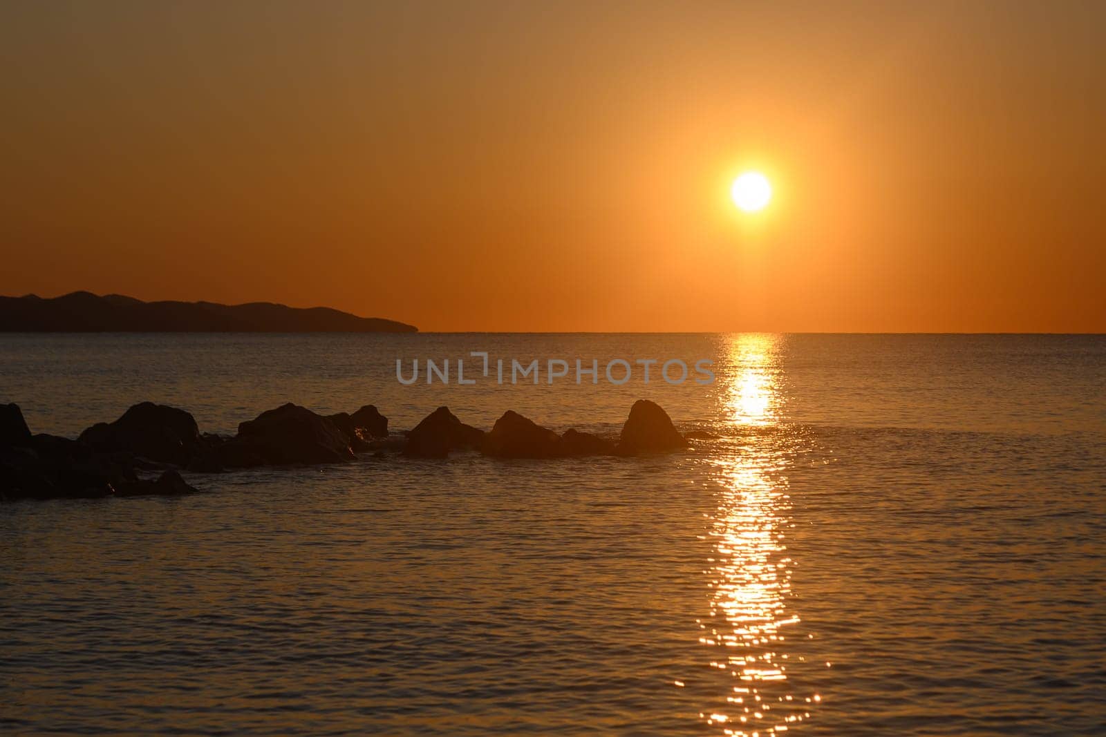 The sun slowly descends in a fiery display, casting warm orange and yellow hues over the serene ocean. Waves gently lap against dark rocks, creating a peaceful ambiance as twilight approaches.