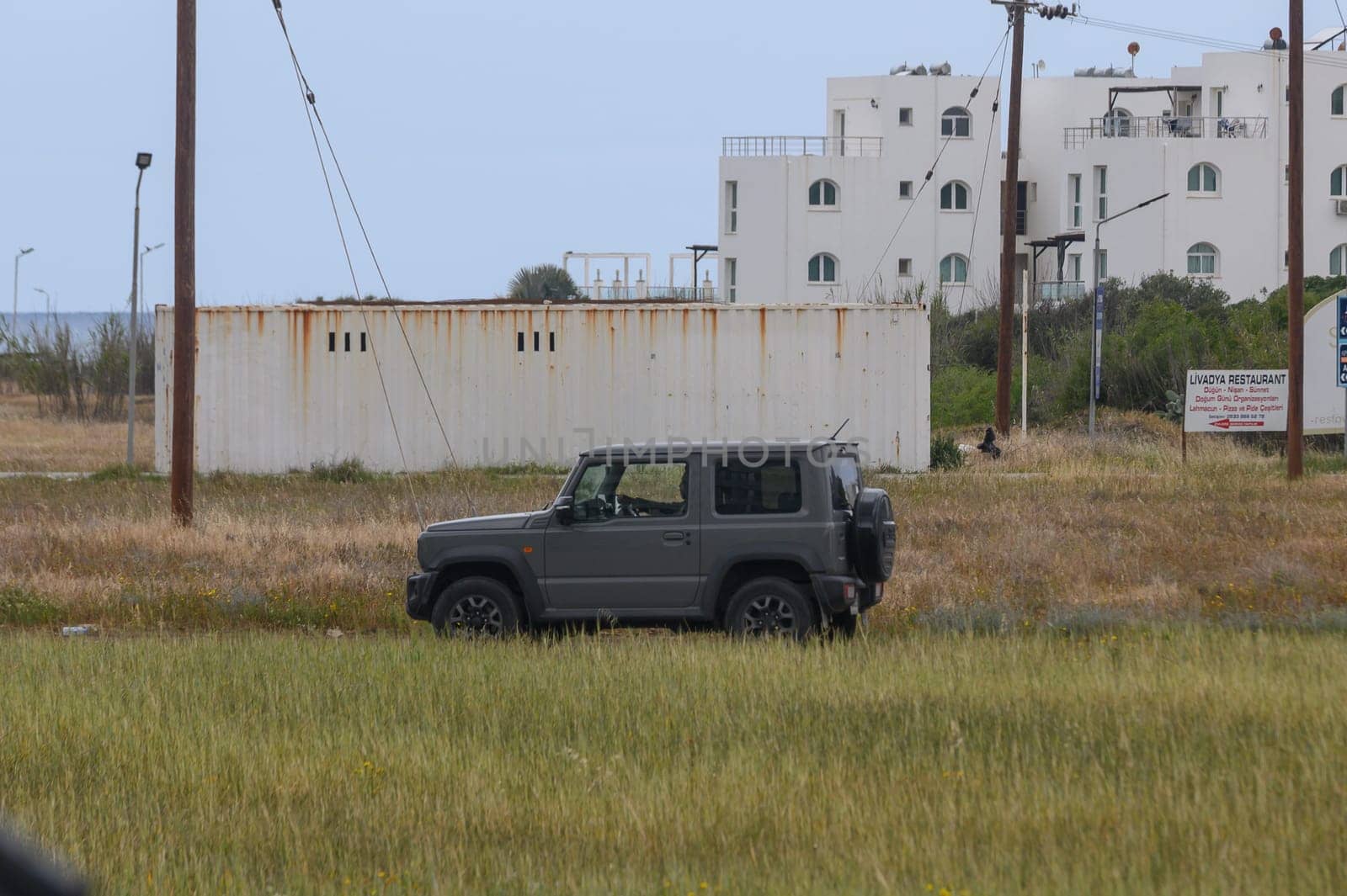 Gaziveren Cyprus -10.04.2025 A rugged vehicle sits in tall grass, overlooking an empty lot marked by an old restaurant and a distant sea. Gray clouds loom overhead, hinting at an untamed adventure ahead.