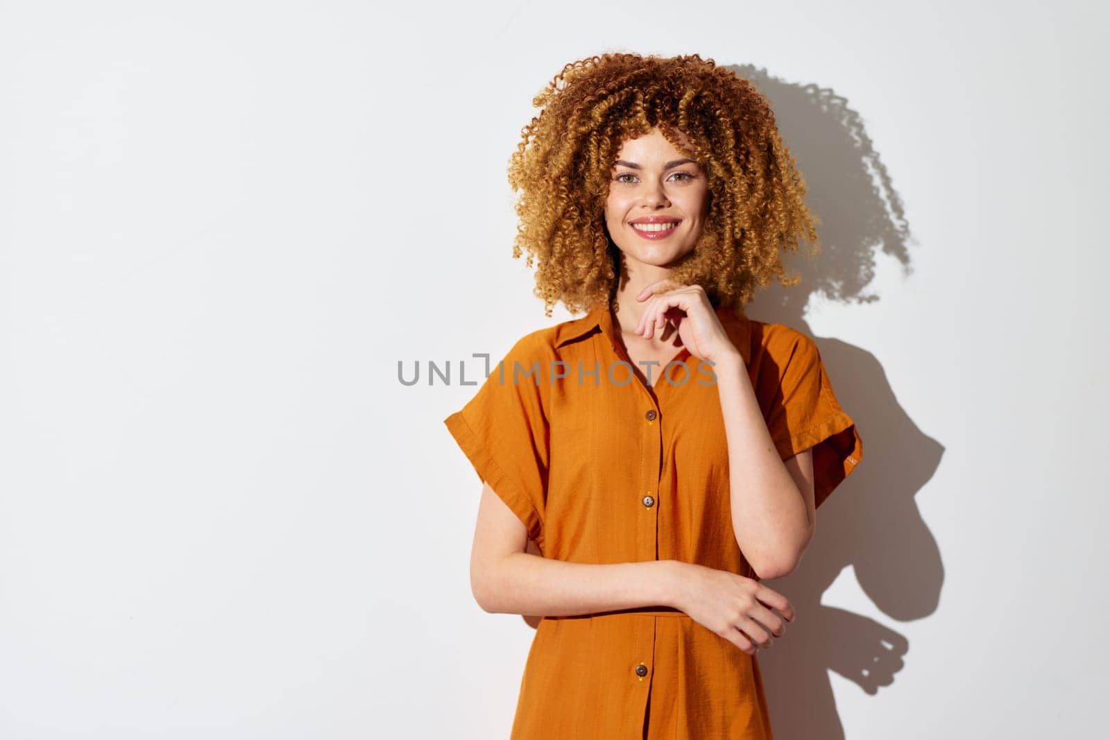 Smiling young woman with voluminous curly hair, dressed in a brown shirt, poses against a plain white backdrop Her cheerful demeanor and stylish look evoke feelings of happiness and positive energy