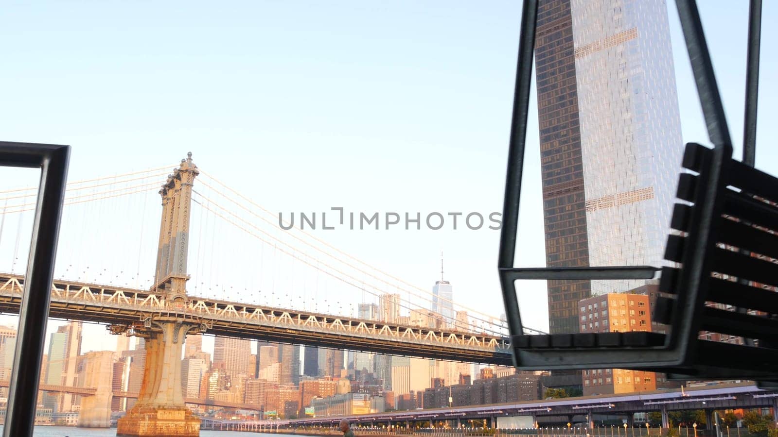 New York City, United States - 7 Sept 2023: Manhattan Bridge to Brooklyn. Pier 35 swings bench, waterfront downtown skyline cityscape. World Trade Center skyscraper, financial district morning sunset.