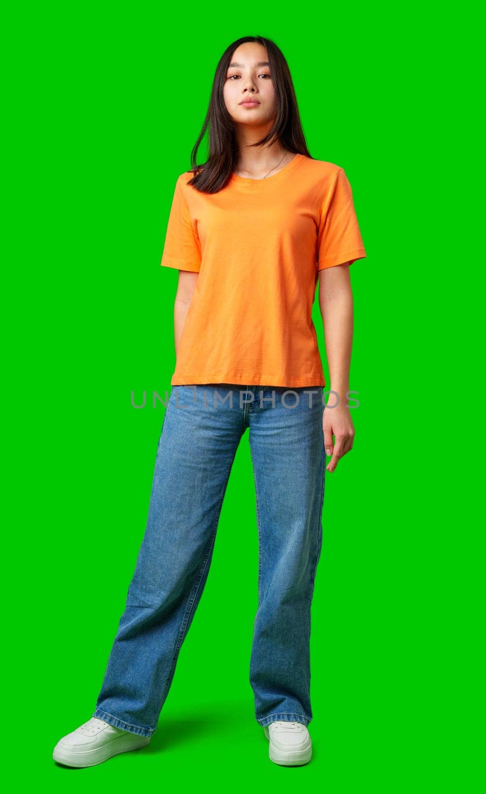A model stands upright wearing a bright orange shirt and blue jeans, showcasing a confident expression. The vivid green backdrop highlights the subject during a studio session.