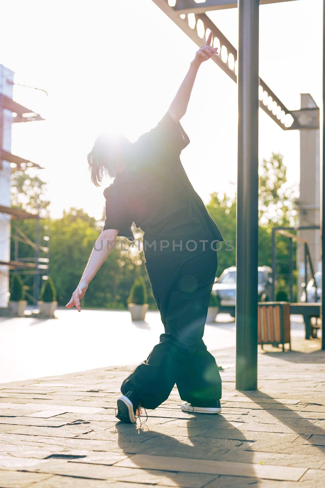 A dancer engages in expressive movement in an urban area as the sun sets, casting a beautiful glow and creating a striking silhouette against the backdrop of trees and buildings.