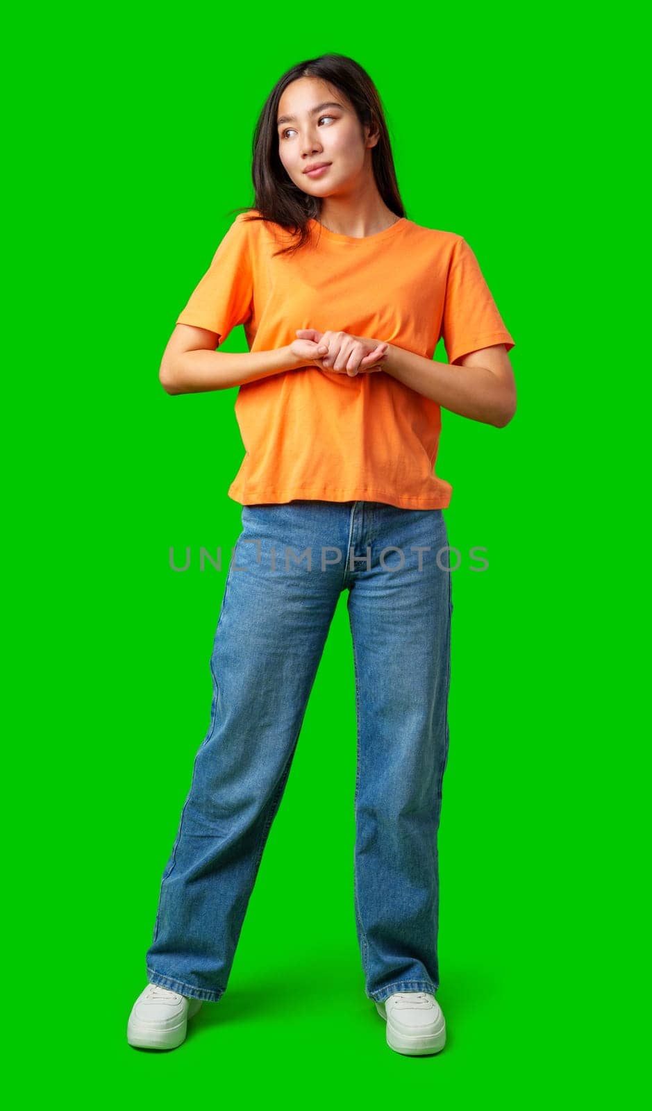A young woman stands with her hands clasped, wearing an orange shirt and blue jeans. The vibrant green background highlights her relaxed yet confident pose.