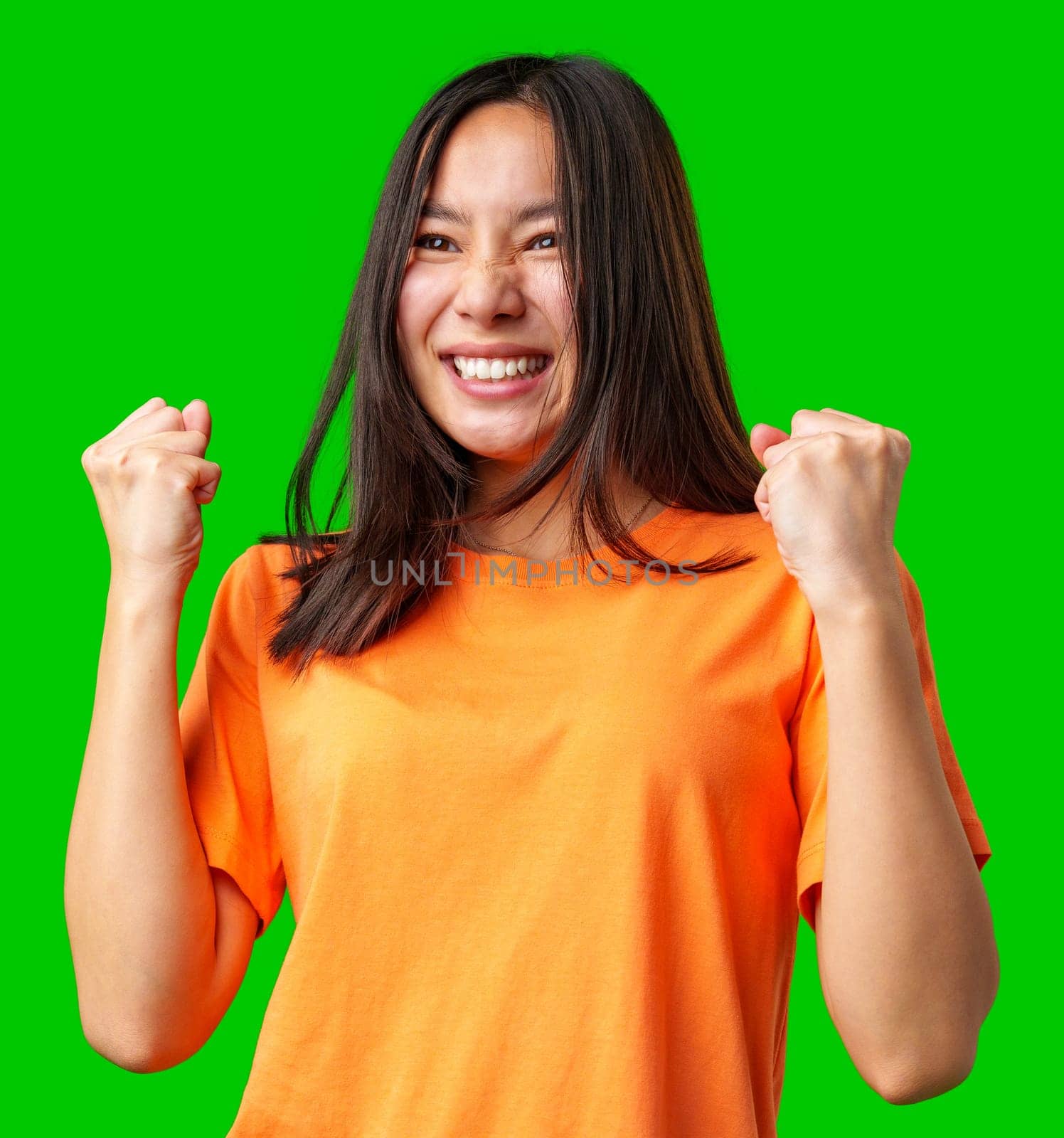 A young woman wearing a vibrant orange shirt is joyfully expressing her excitement, with her fists raised in celebration. The bright green background enhances the lively atmosphere.