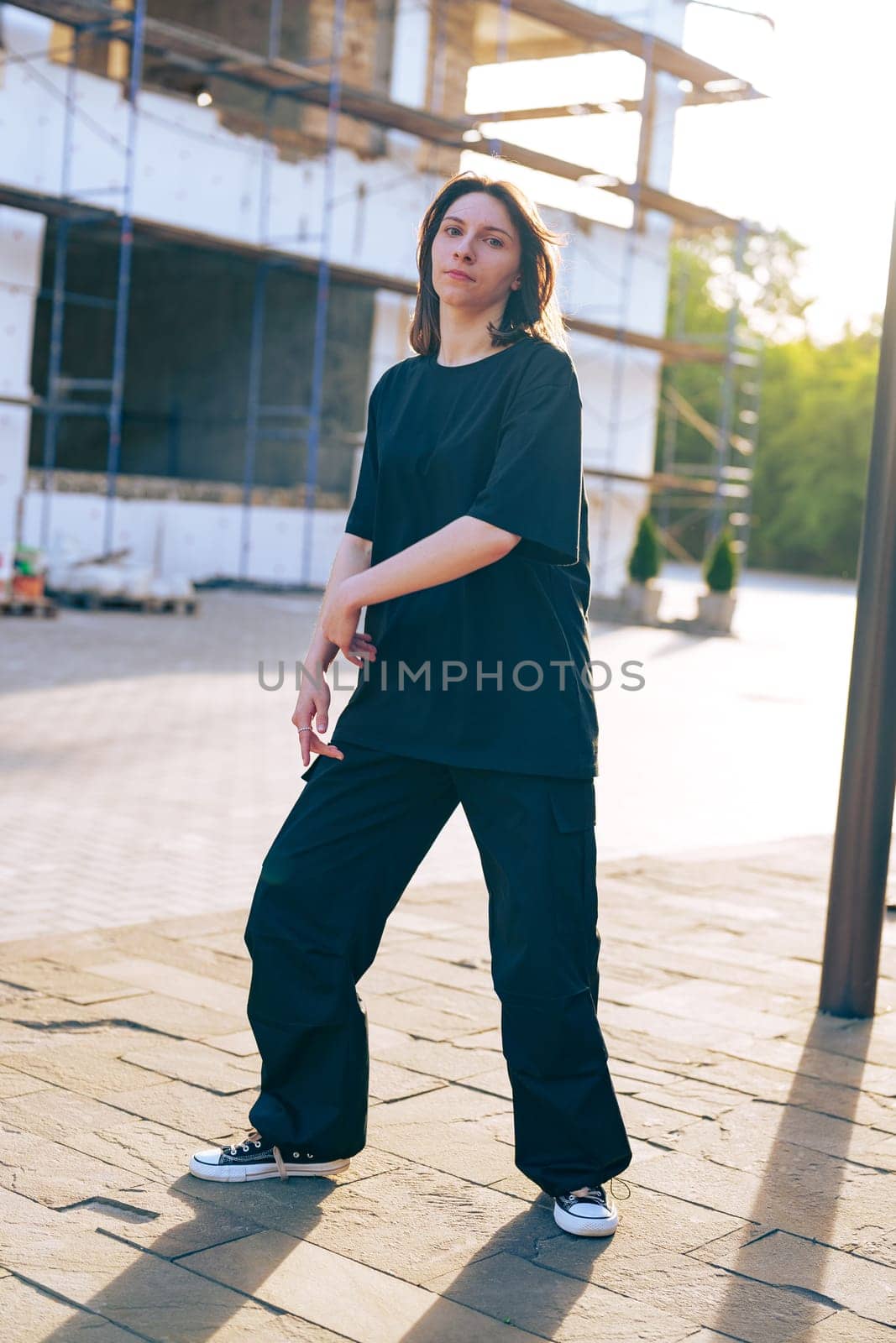 A young woman wearing a loose black shirt and wide-legged pants strikes a pose in front of a construction site, basking in the warm light of the setting sun.