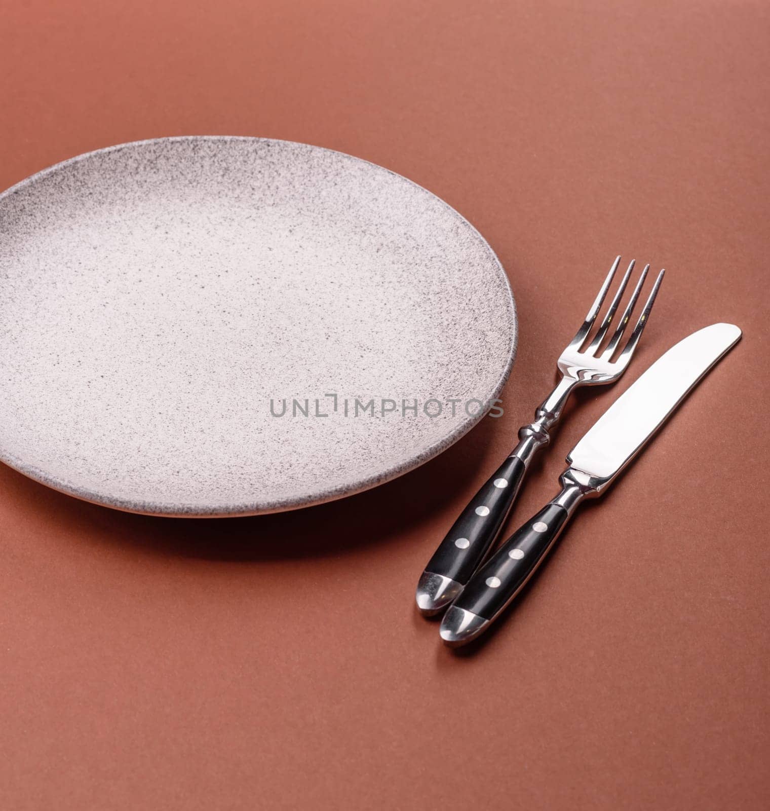 Empty ceramic plate on a monochrome brown background. Empty plate with cutlery on a dining table