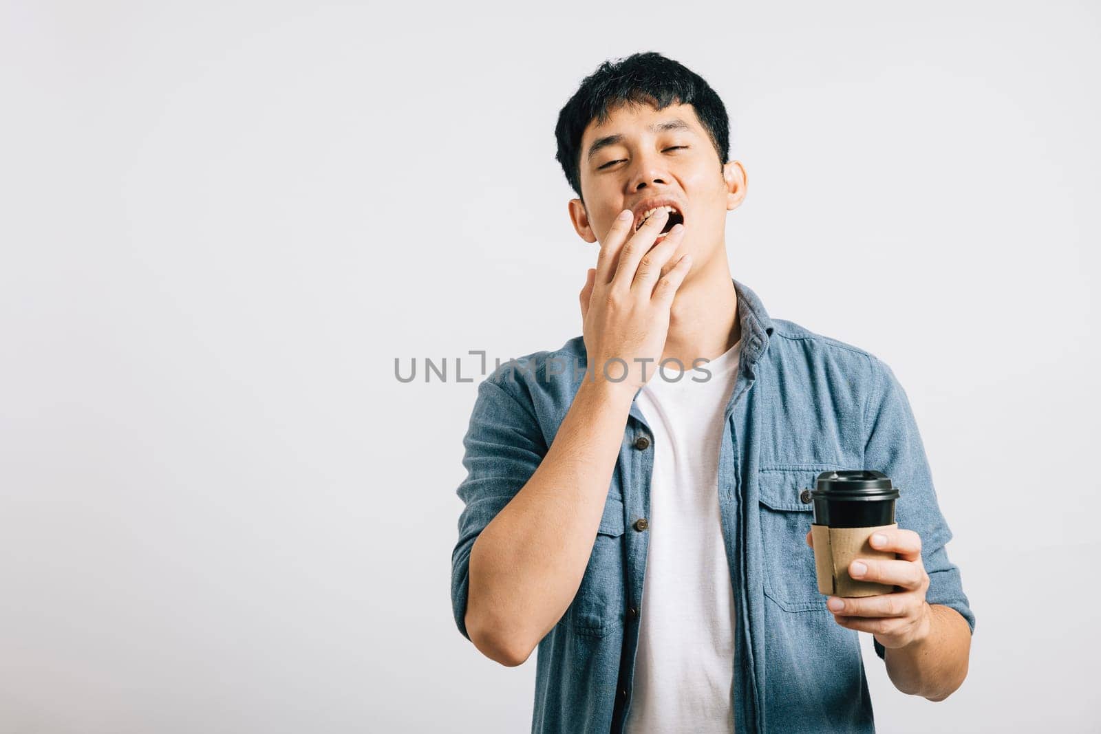 A young man, overwhelmed by fatigue, yawns with his eyes closed, coffee cup in hand. Studio shot isolated on white, his tired expression tells a relatable story. Copyspace included.