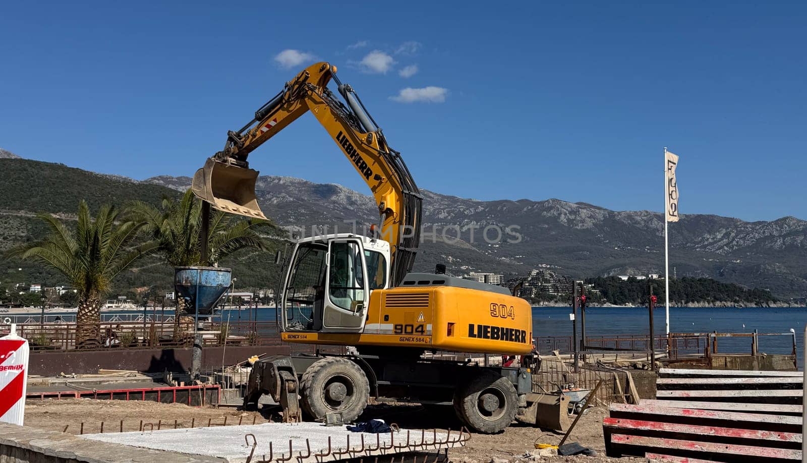 Budva,Montenegro-April 11,2025:A yellow Liebherr excavator working near the coast filling a concrete crater at a coastal construction site with palm trees and a COCO cafe flag. High quality photo