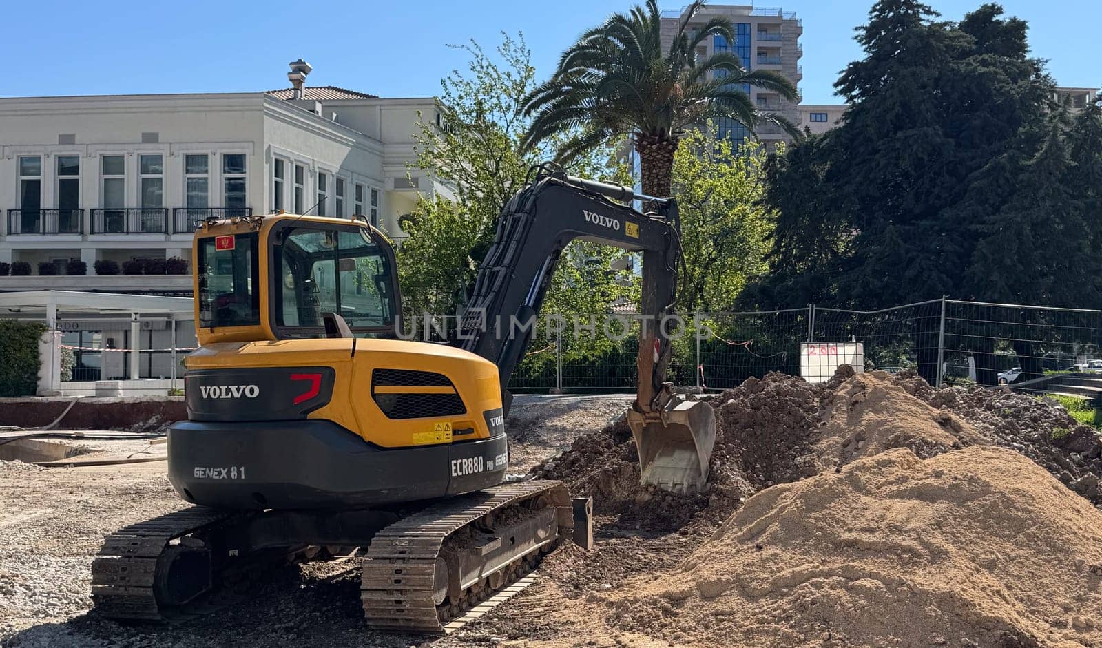 Budva,Montenegro-April 11,2025: Yellow and black Volvo excavator digging soil at a city construction site surrounded by buildings and palm trees.