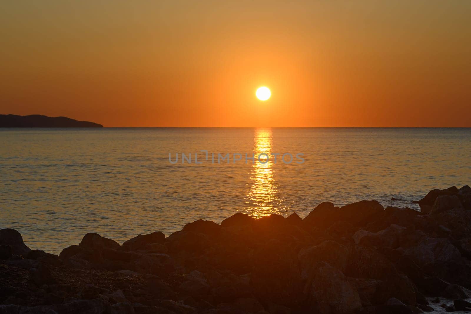 As the sun sets on the horizon, hues of gold and orange illuminate the calm sea, creating a peaceful ambiance. Silhouetted rocks frame this picturesque moment, inviting tranquility.