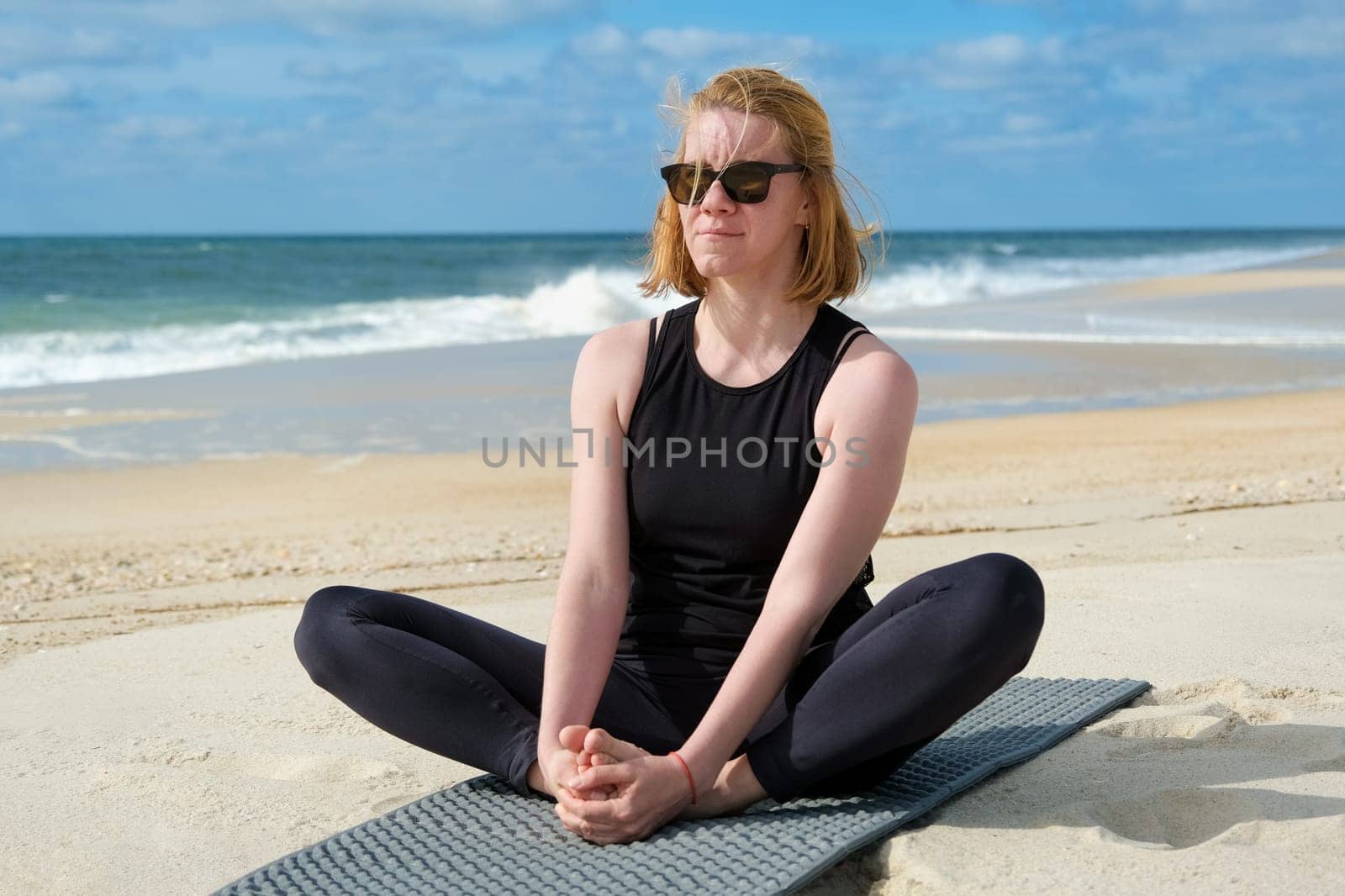 Woman practicing yoga on sandy beach, sitting in butterfly pose on mat, embracing mindfulness, relaxation, and wellness with ocean waves and blue sky background.