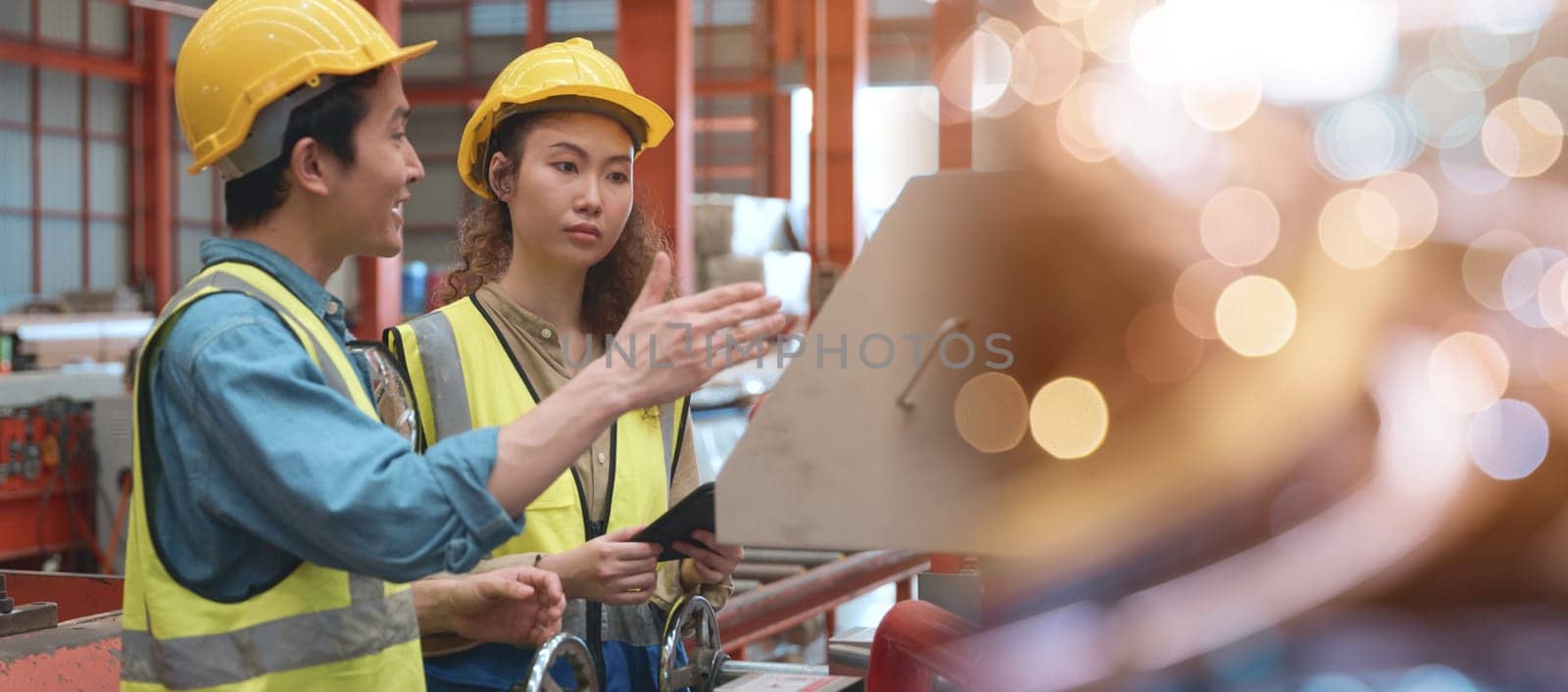 Industrial engineer man and worker operation woman training work on metal sheet ironing machine, two engineering wear safety helmet they talking together about machinery in factory manufacture