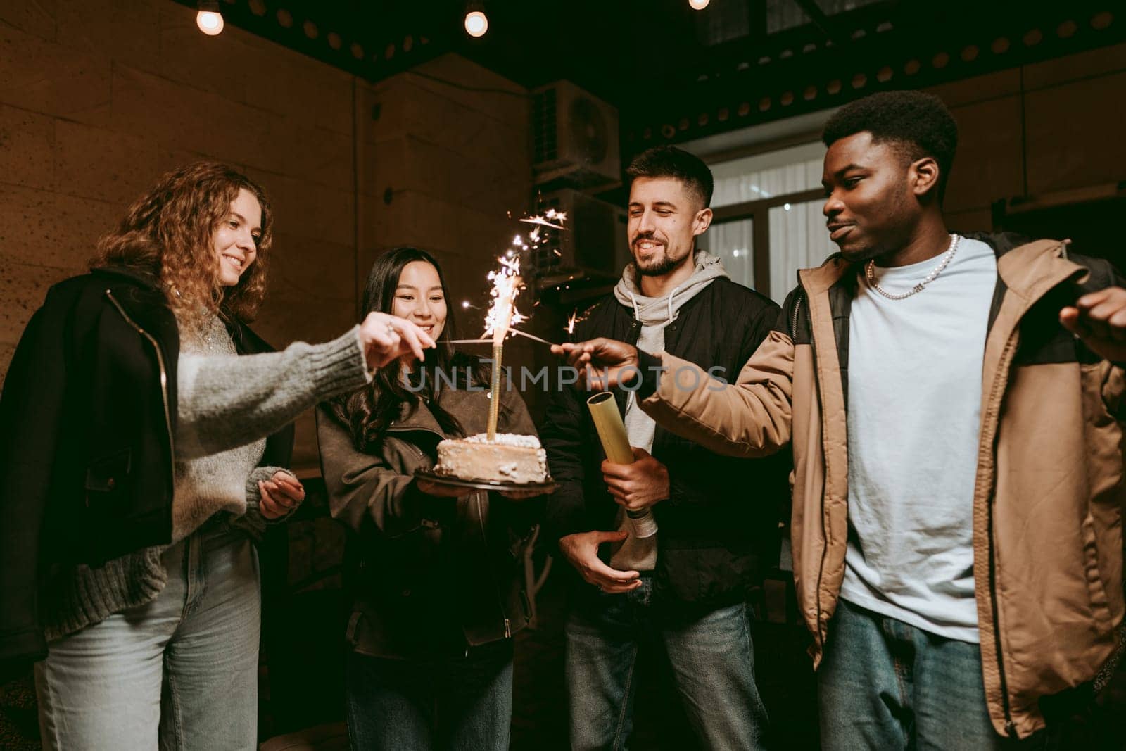 A group of four friends gather around a cake with sparklers during a birthday celebration in a warm indoor environment filled with festive lights and joyful laughter.