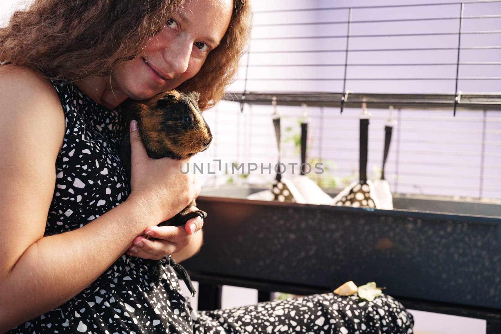 A young girl smiles as she gently cradles a guinea pig close to her chest. She sits comfortably beside a pet cage, creating a warm atmosphere for bonding with her furry friend.