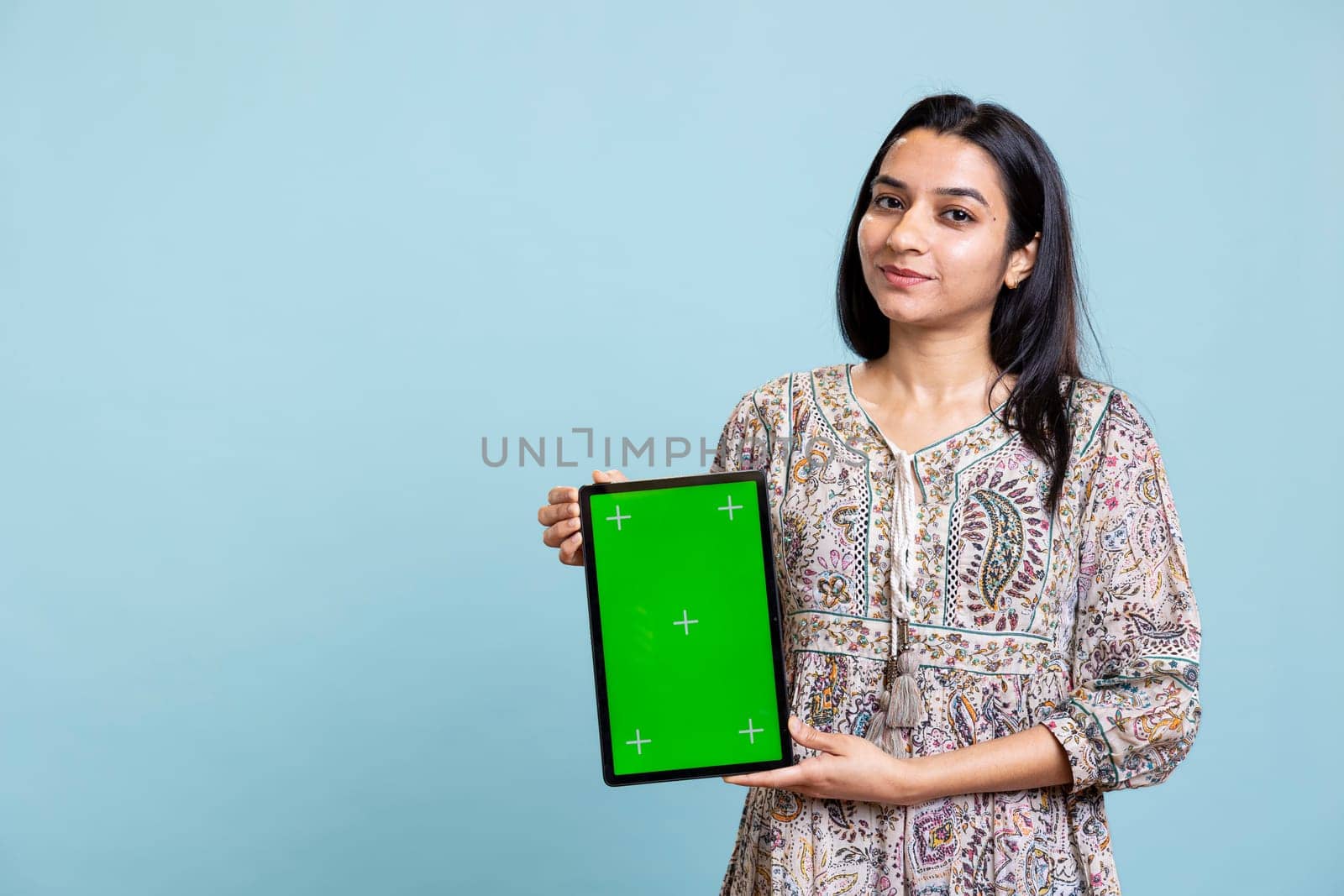 Indian girl smiling and showing isolated copy space screen in front of camera, creating an advertisement for a commercial against blue background. Young joyful adult holding device in studio.