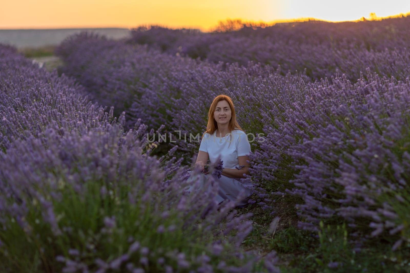 A woman sits in a field of lavender flowers. The flowers are purple and the sky is orange