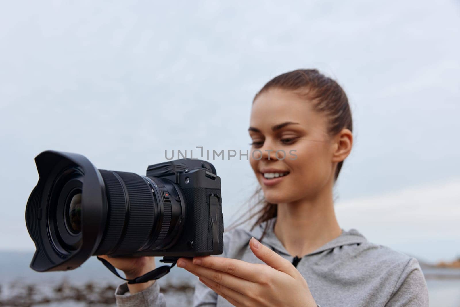 A cheerful young woman wearing casual gray clothing holds a camera at the beach, surrounded by a calm ocean landscape Capturing the essence of creativity and passion for photography