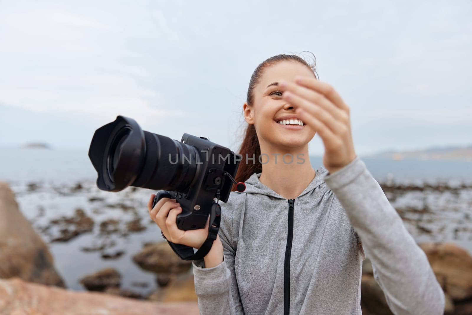 A smiling young woman, dressed in casual athletic wear, joyfully takes photos by the beach, showcasing her love for photography and the outdoors