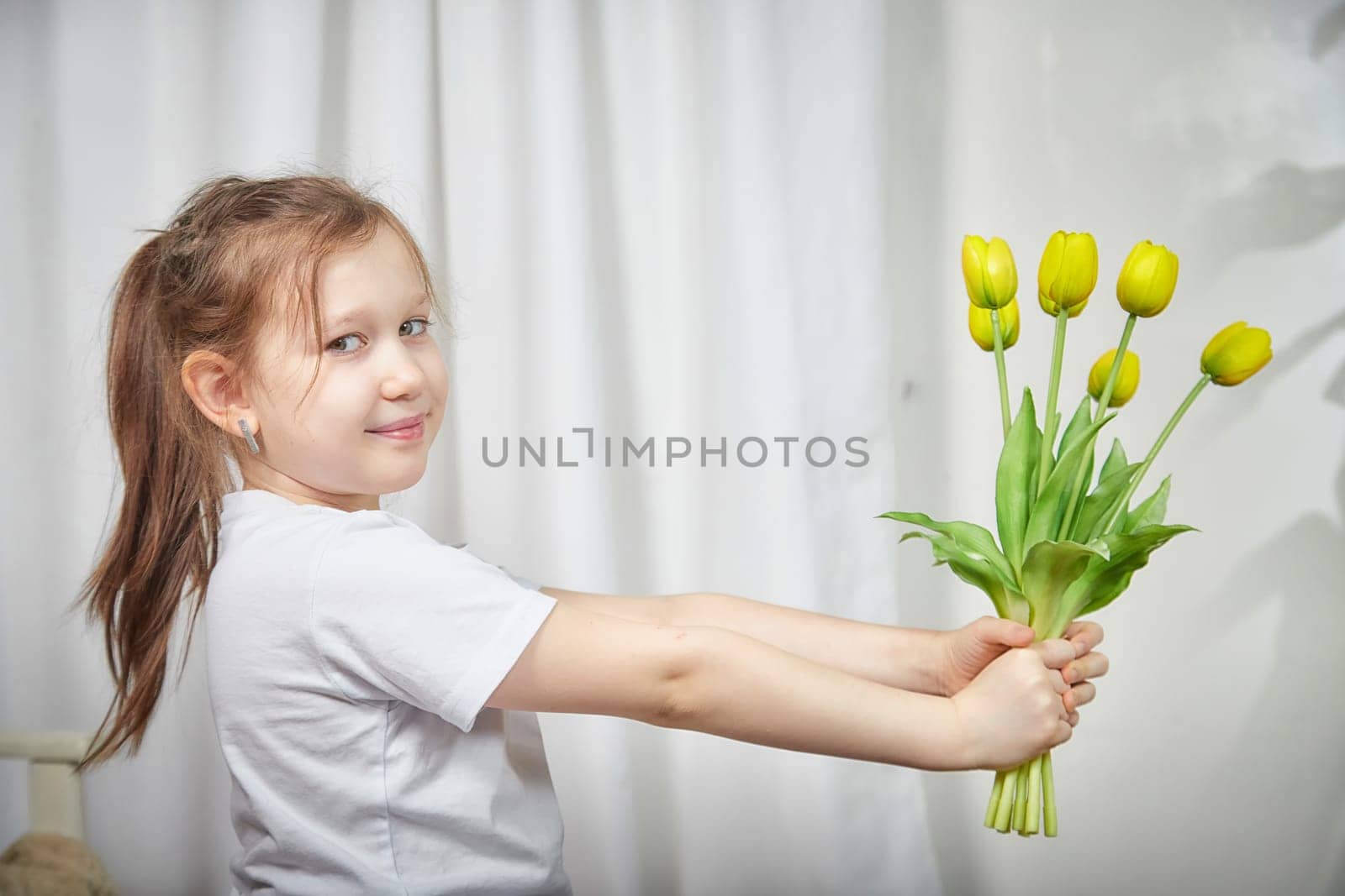 A young girl is happily presenting a bouquet of yellow tulips while standing in a bright room with soft curtains.