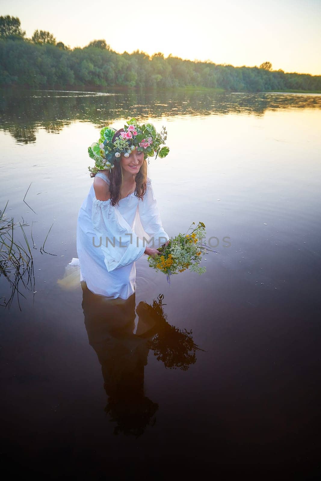 Slavic plump chubby girl in long white dress on the feast of Ivan Kupala with flowers and water in a river or lake on summer evening