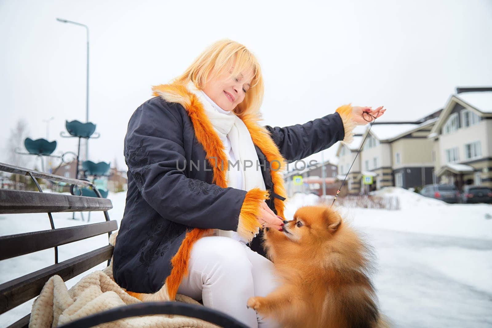 A middle-aged woman with blonde hair enjoys a winter walk, playfully interacting with her fluffy Pomeranian in the snow.