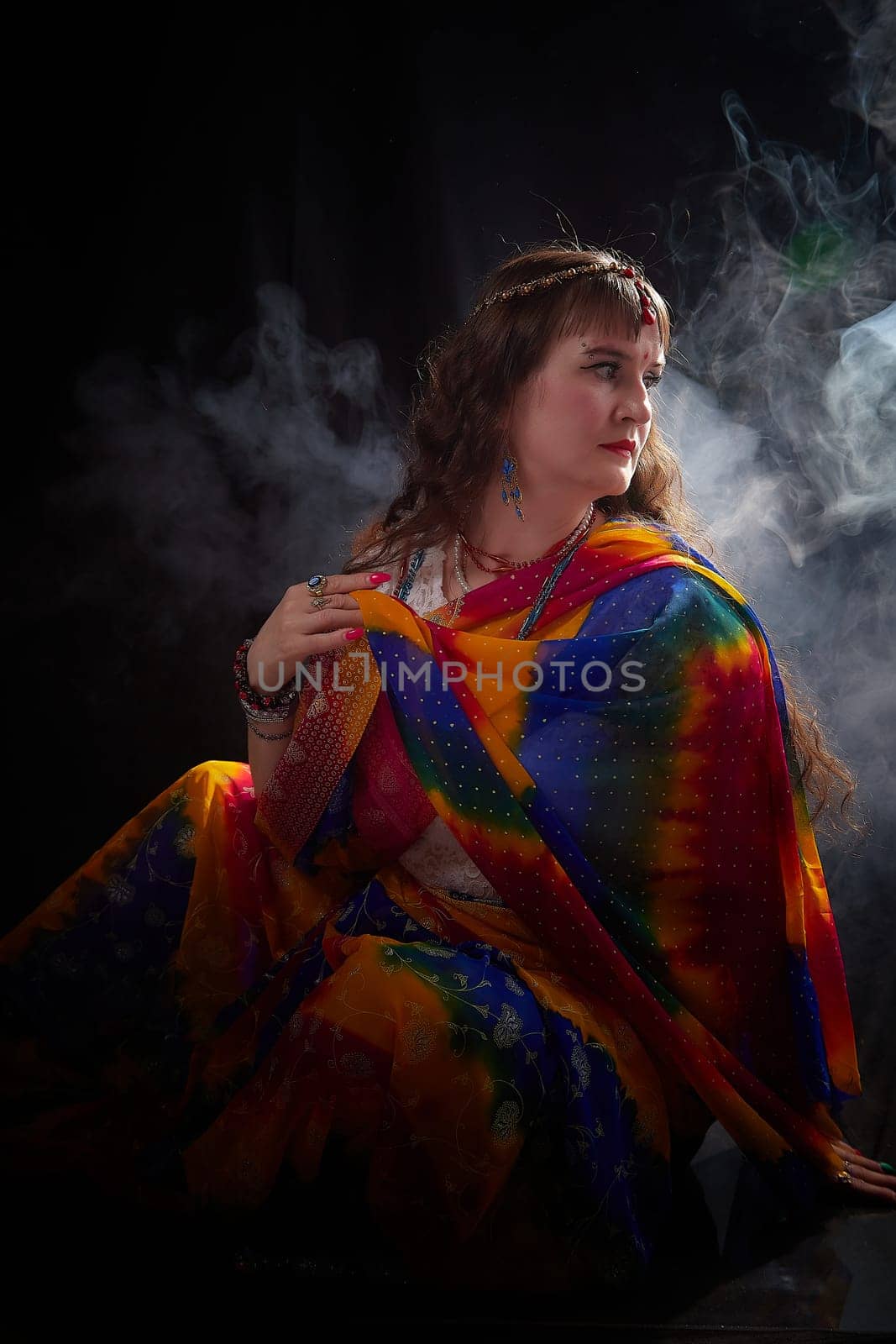 A mature blonde woman poses gracefully in a colorful sari, surrounded by a smoky atmosphere that enhances the artistic vibe.