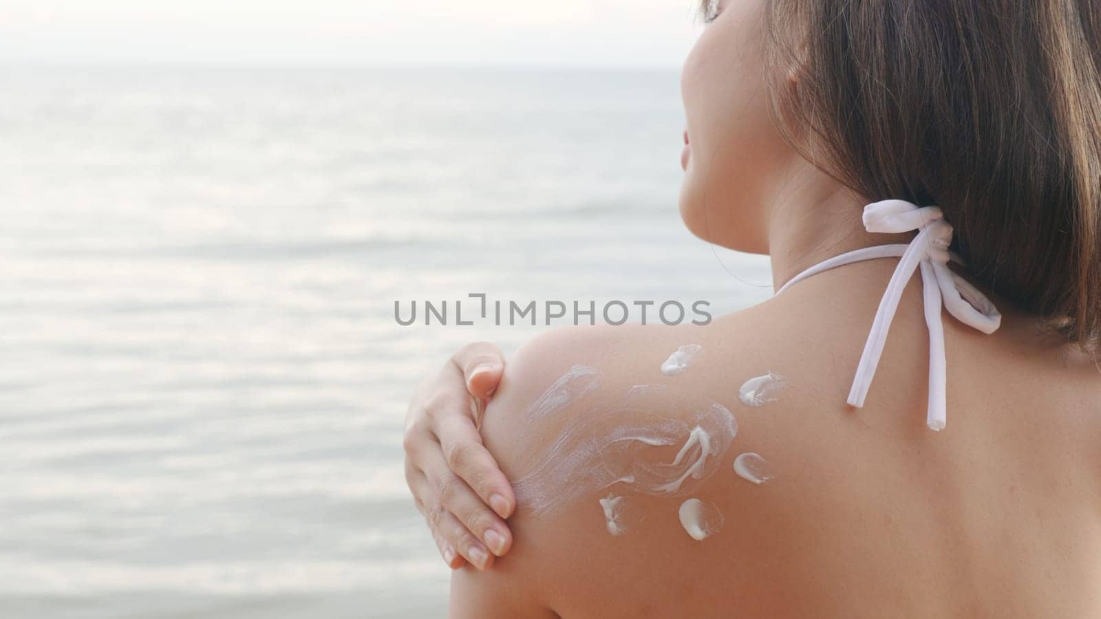 Beautiful Asian woman applying sun protection cream on tanned shoulder on the beach, sun Cream, Close up female in bikini using sunscreen lotion on back body before sunbathing