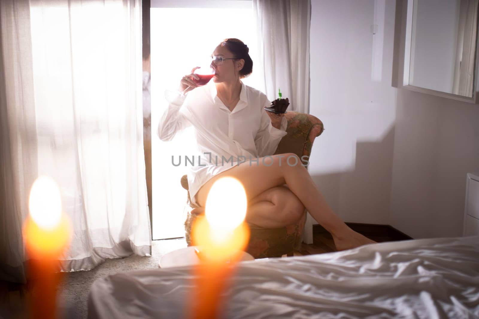 Woman enjoying wine and cake while sitting in armchair near window in hotel room