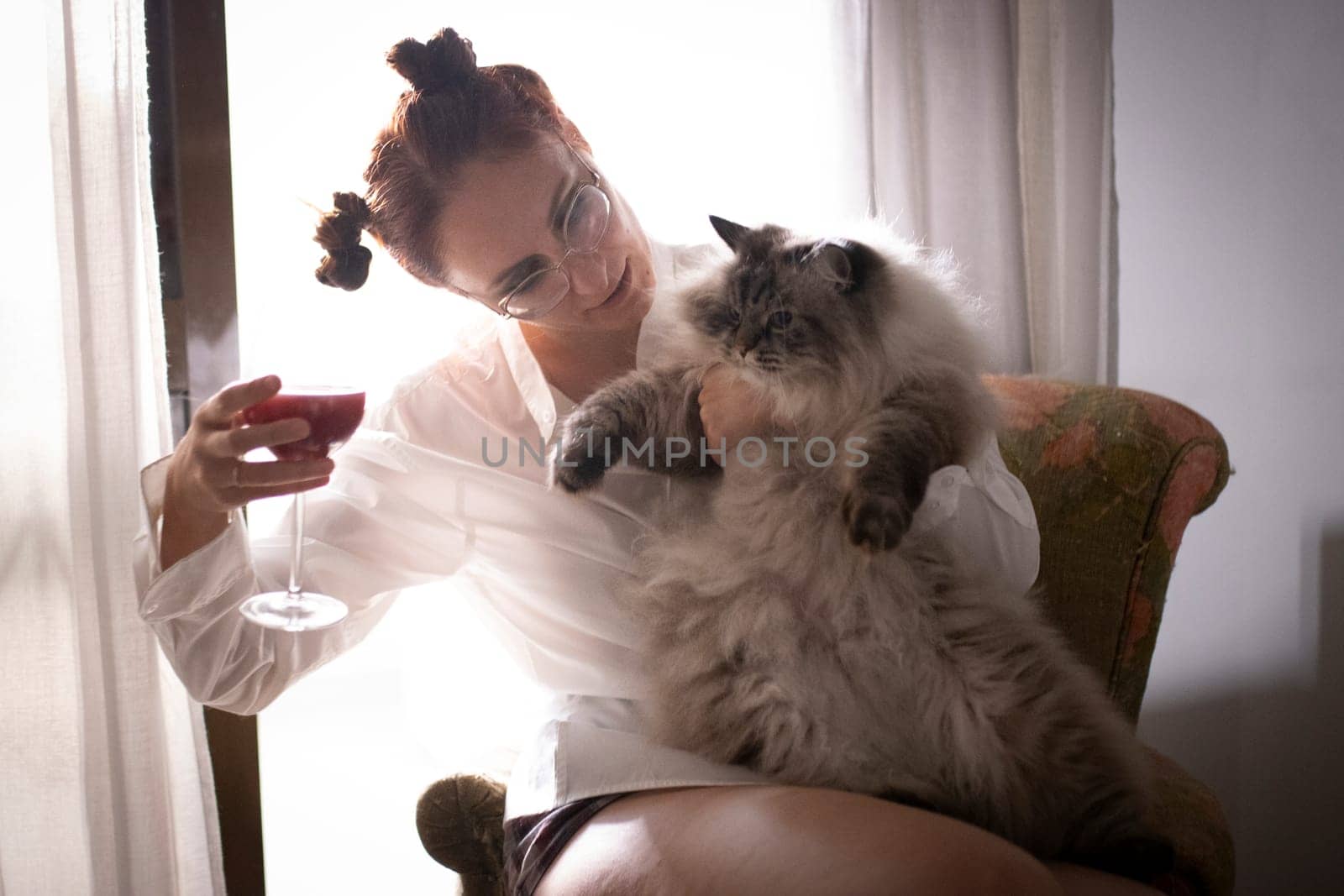 Young woman enjoying a glass of red wine while holding her fluffy cat, relaxing by the window in natural light