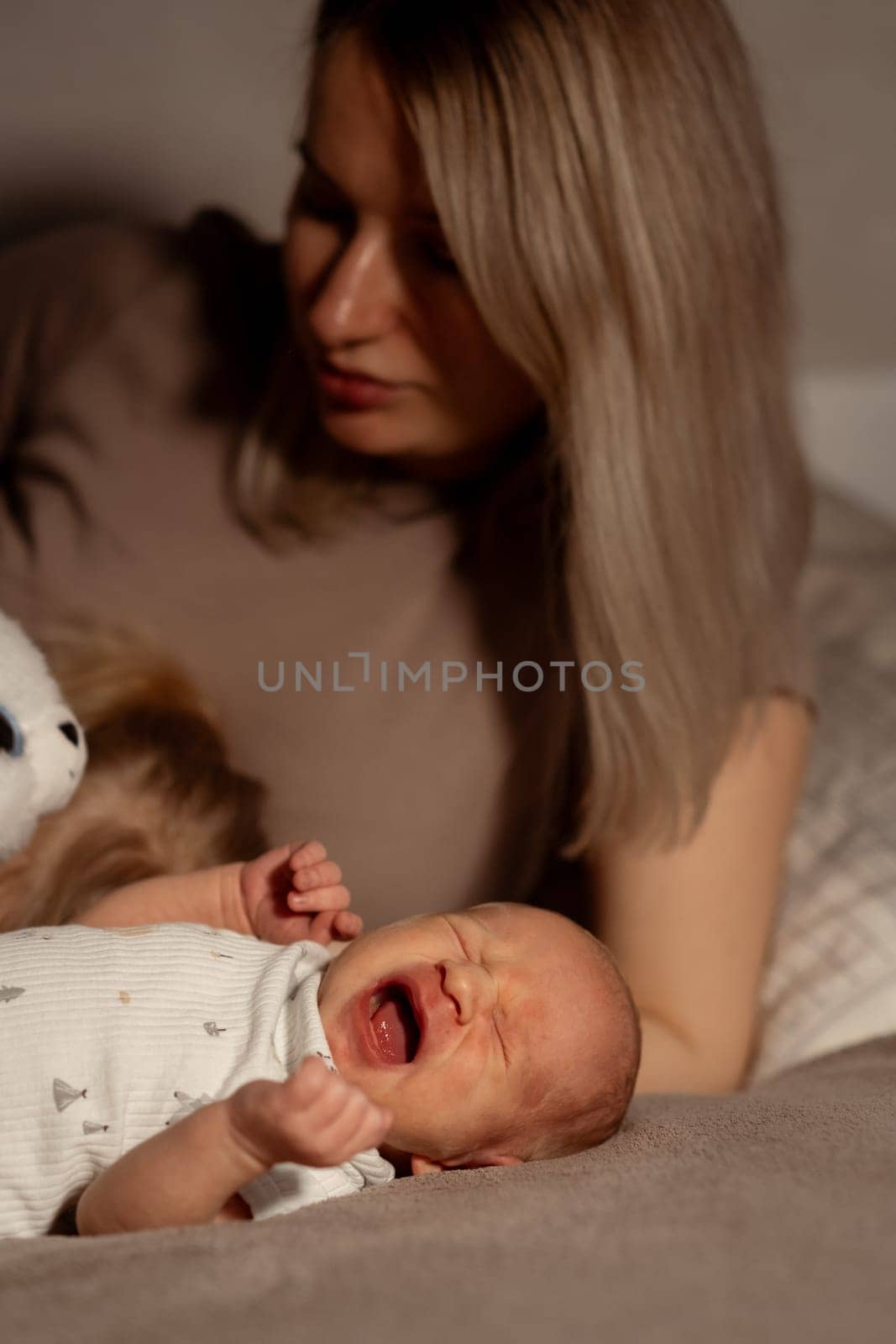 In a serene bedroom, a mother tenderly soothes her crying baby, holding a soft toy while creating a nurturing atmosphere in the evening light.