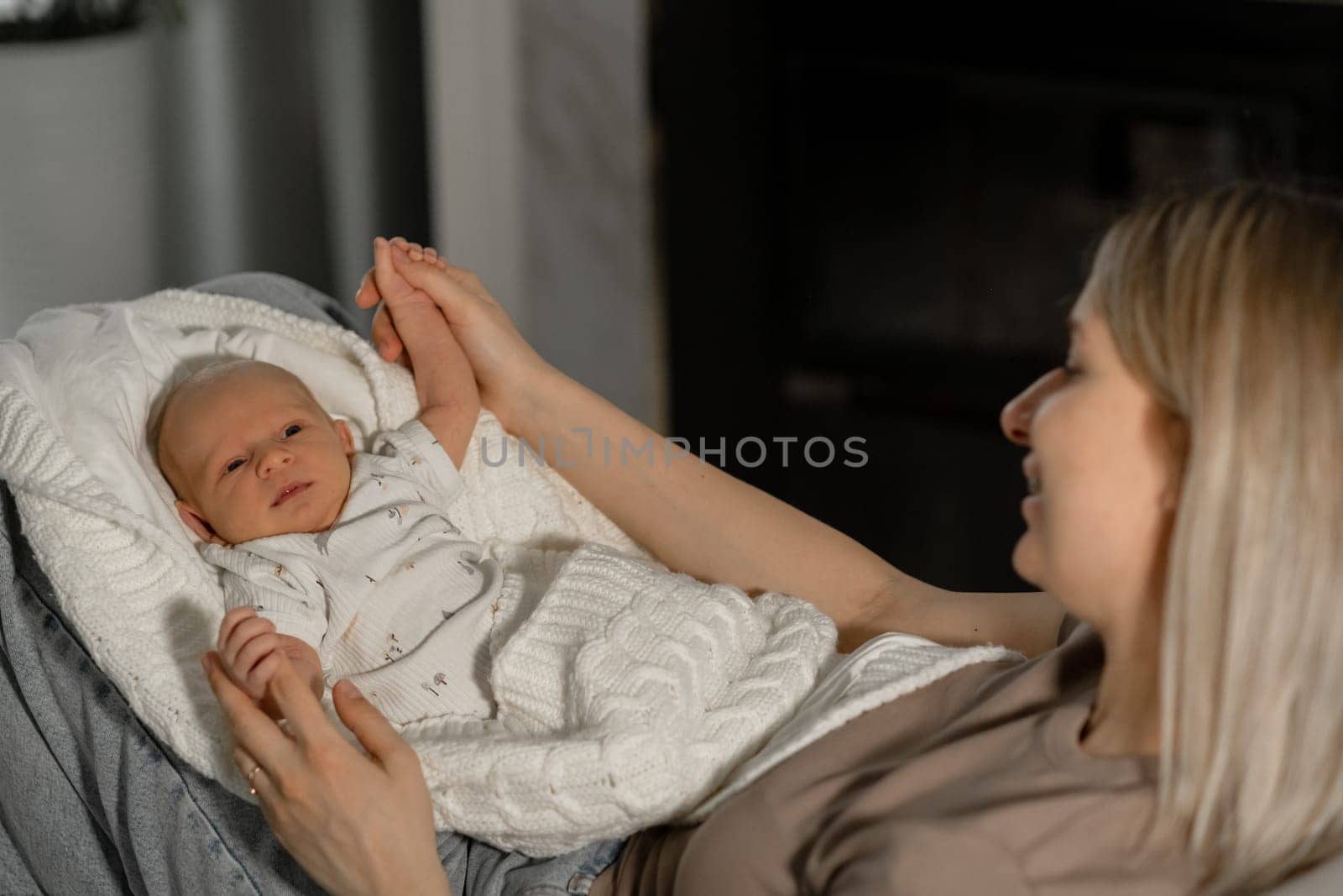 A mother lovingly interacts with her baby, cradling the child in a blanket while sitting comfortably indoors, creating a warm atmosphere.