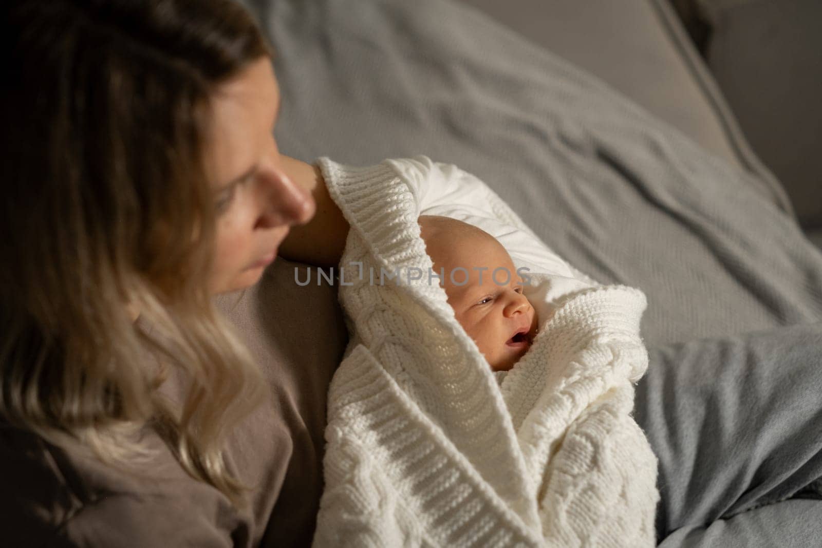 In a calm indoor setting, a mother holds her wrapped newborn, both embracing a peaceful moment of bonding on a soft bed at dusk.