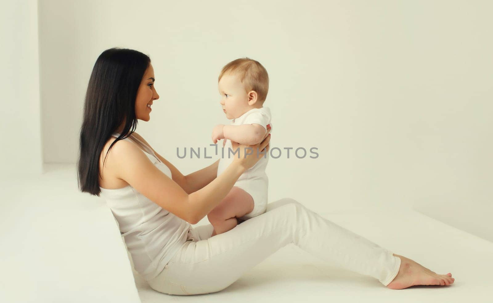 Happy smiling young mother playing with cute baby sitting together in white room at home