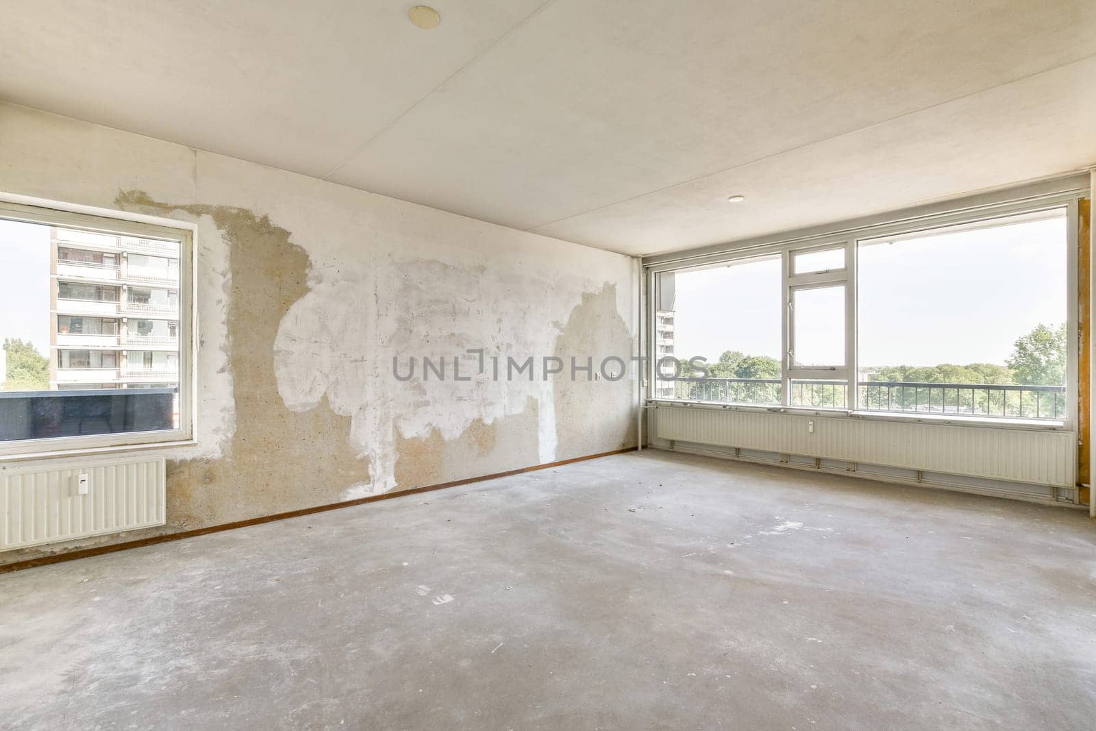An empty room showcasing unfinished walls and expansive windows that offer a view of the outdoors. The floor is bare cement, highlighting the minimalistic space.