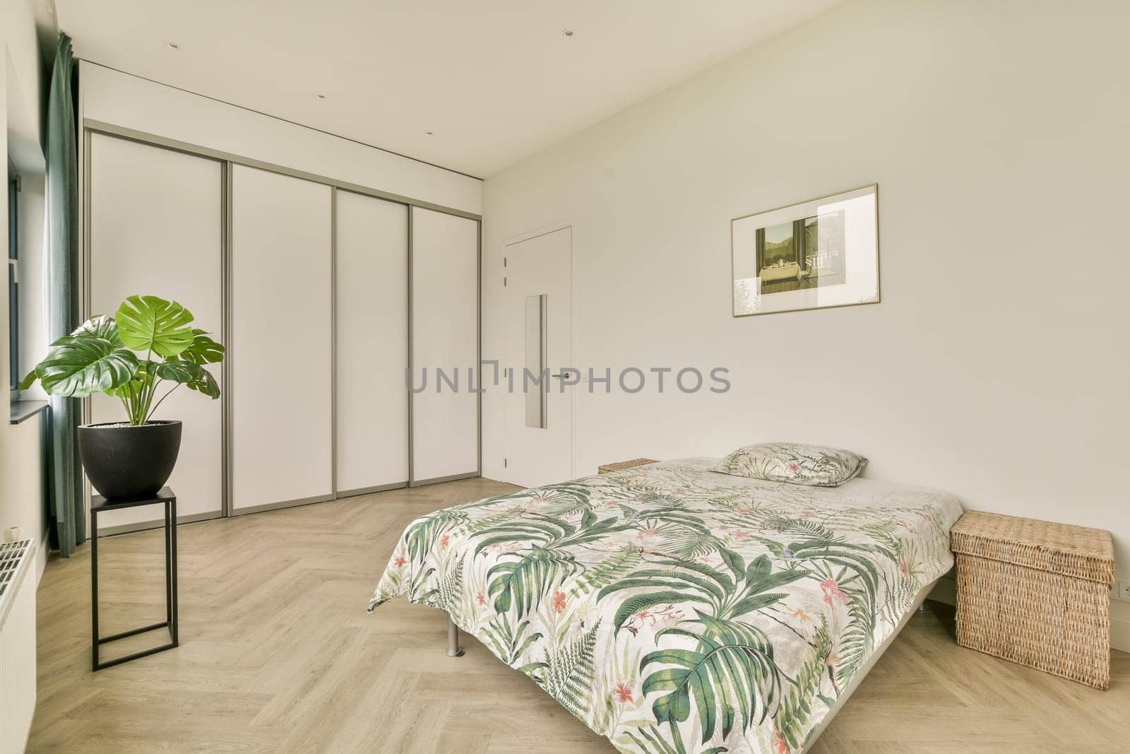 A stylish minimalist bedroom featuring a floral tropical patterned bedspread, large windows, and a decorative potted plant, emphasizing comfort and elegance.