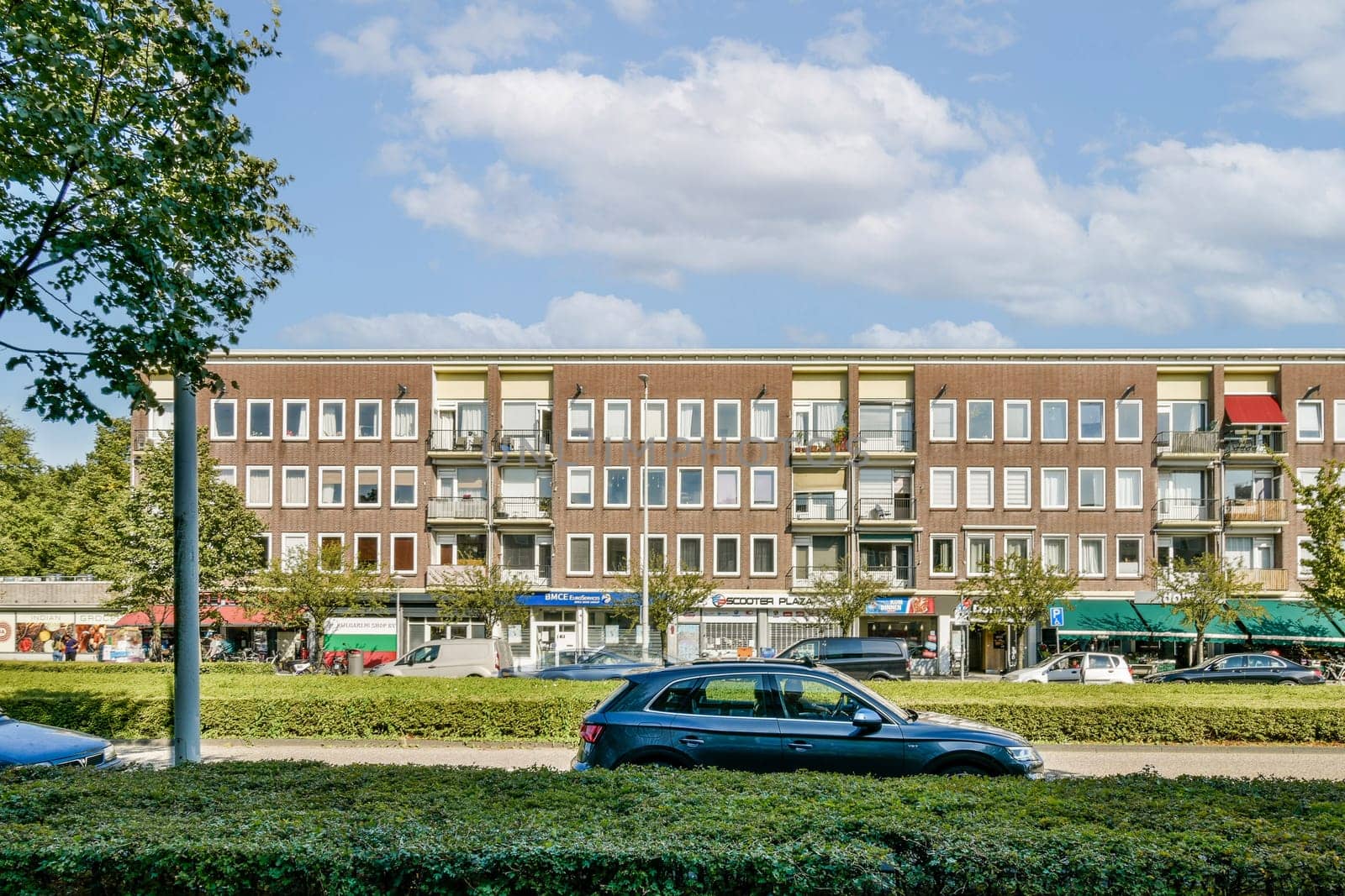 A contemporary urban building featuring multiple shops at the ground level and residential units above, surrounded by greenery and a clear blue sky.